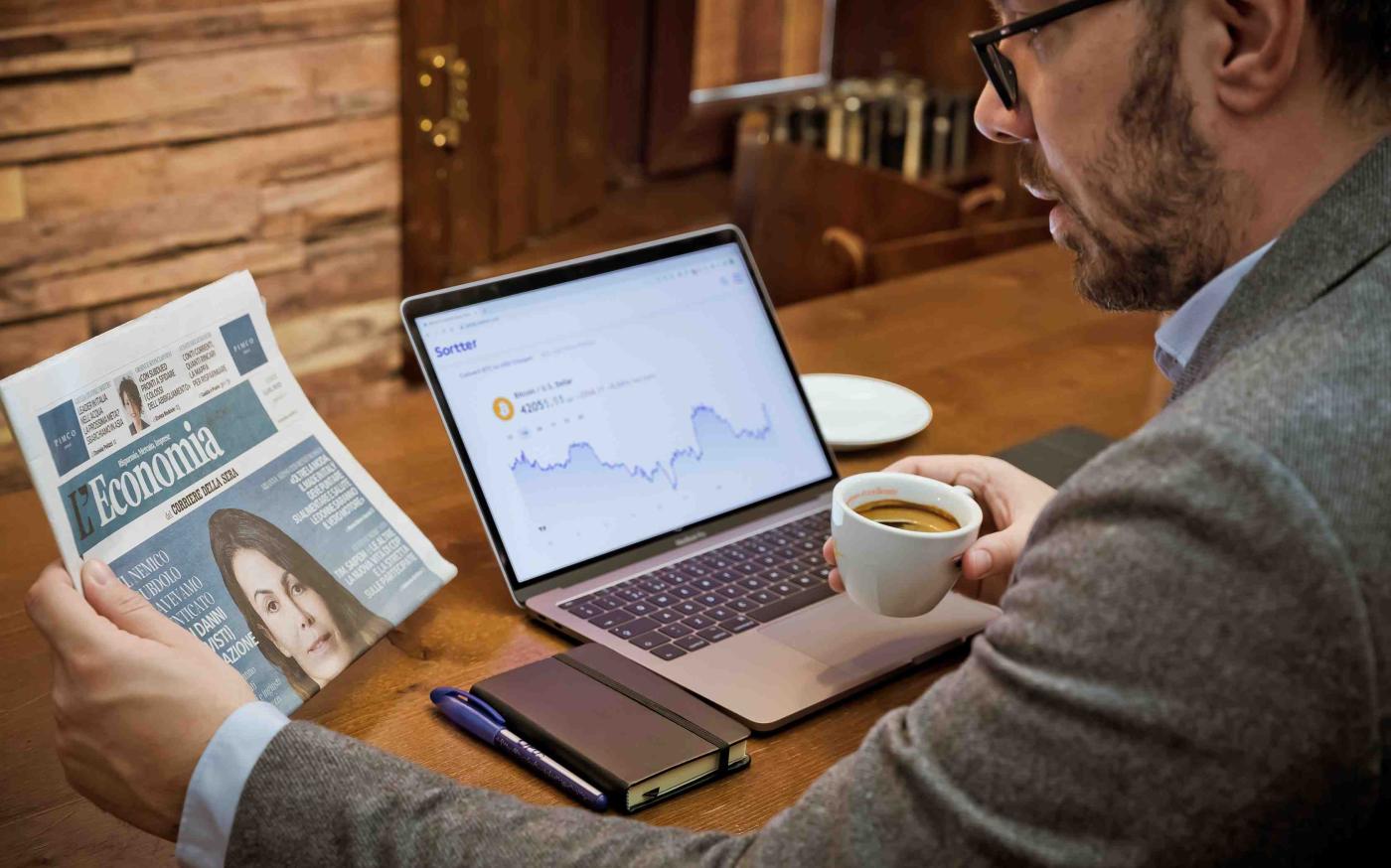 Man reading newspaper next to laptop and phone